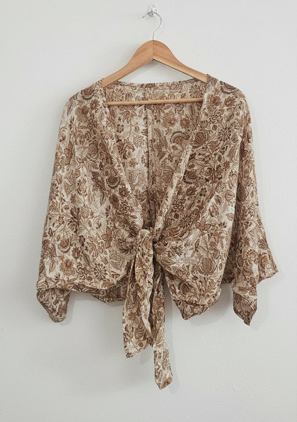 A lightweight beige and brown paisley print wrap top hanging on a wooden hanger against a paneled wall.