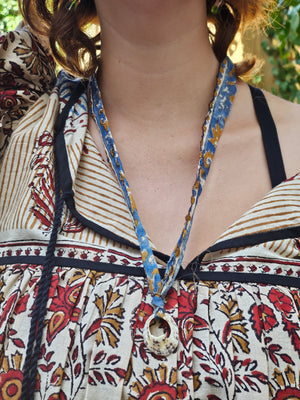 A close-up of a person wearing a bohemian-style necklace with a blue and yellow patterned fabric ribbon and a cowrie shell pendant, paired with a matching patterned blouse.