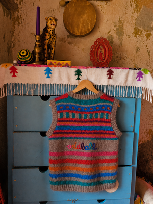 A colorful hand-knit sweater vest hangs on a wooden hanger from a distressed blue dresser in a rustic room. The vest features horizontal stripes of blue, teal, orange, pink, and gray, with a patterned yoke and the word 'oddball' knitted on the chest.