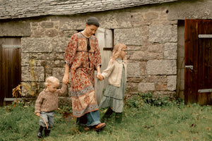 A woman and two children walking outdoors in front of a rustic stone building, wearing earthy-toned vintage clothing. The woman wears a long patterned dress and dark hat, holding hands with the children.