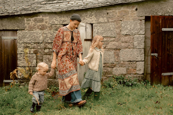 A woman and two children walking outdoors in front of a rustic stone building, wearing earthy-toned vintage clothing. The woman wears a long patterned dress and dark hat, holding hands with the children.